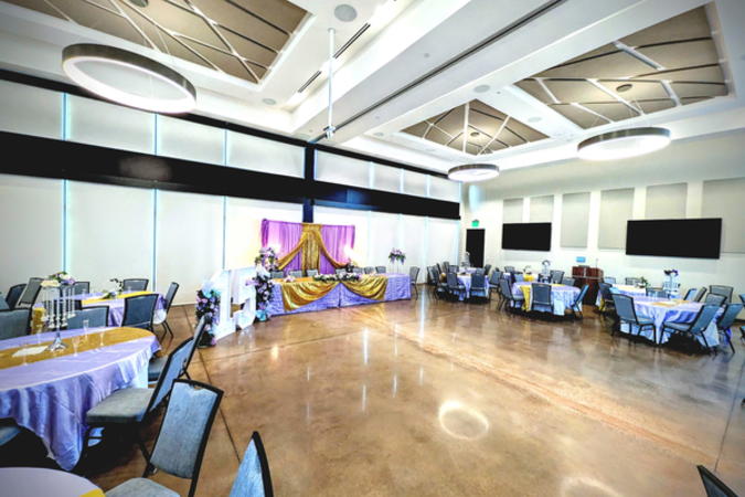 Angle of the event center showing the brown glossy floor, wall monitors, round tables, and large round light fixtures hanging from the ceiling