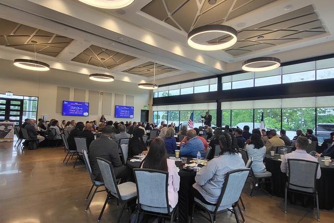 Business event happening inside the event center. Catered food is offered in the left of the photo, next to the catering kitchen. Round tables are filled with people eating and watching a presentation.
